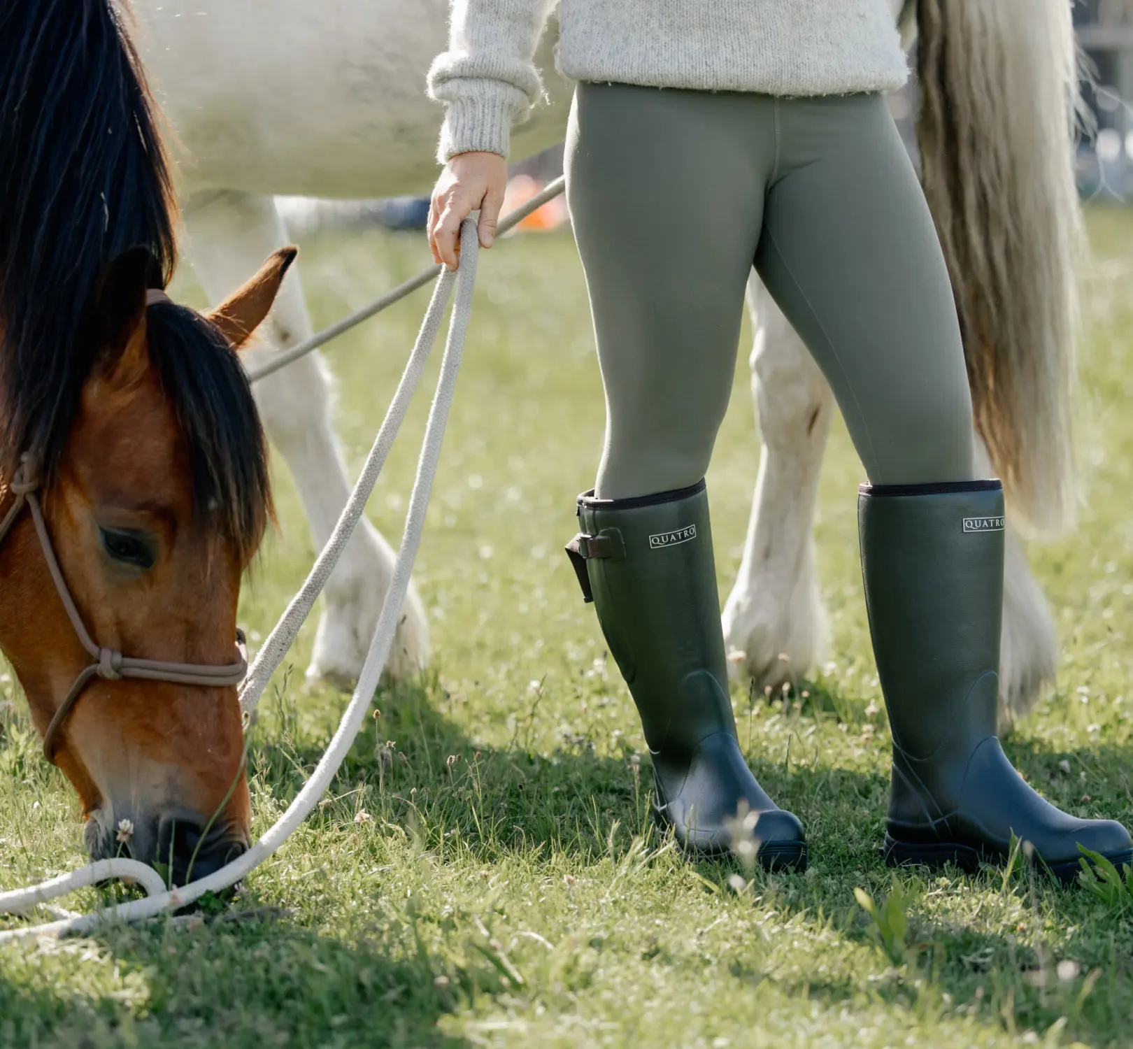 A person in green rubber boots and leggings holds a lead rope as a brown horse with a white blaze grazes on grass; a second white horse stands in the background.