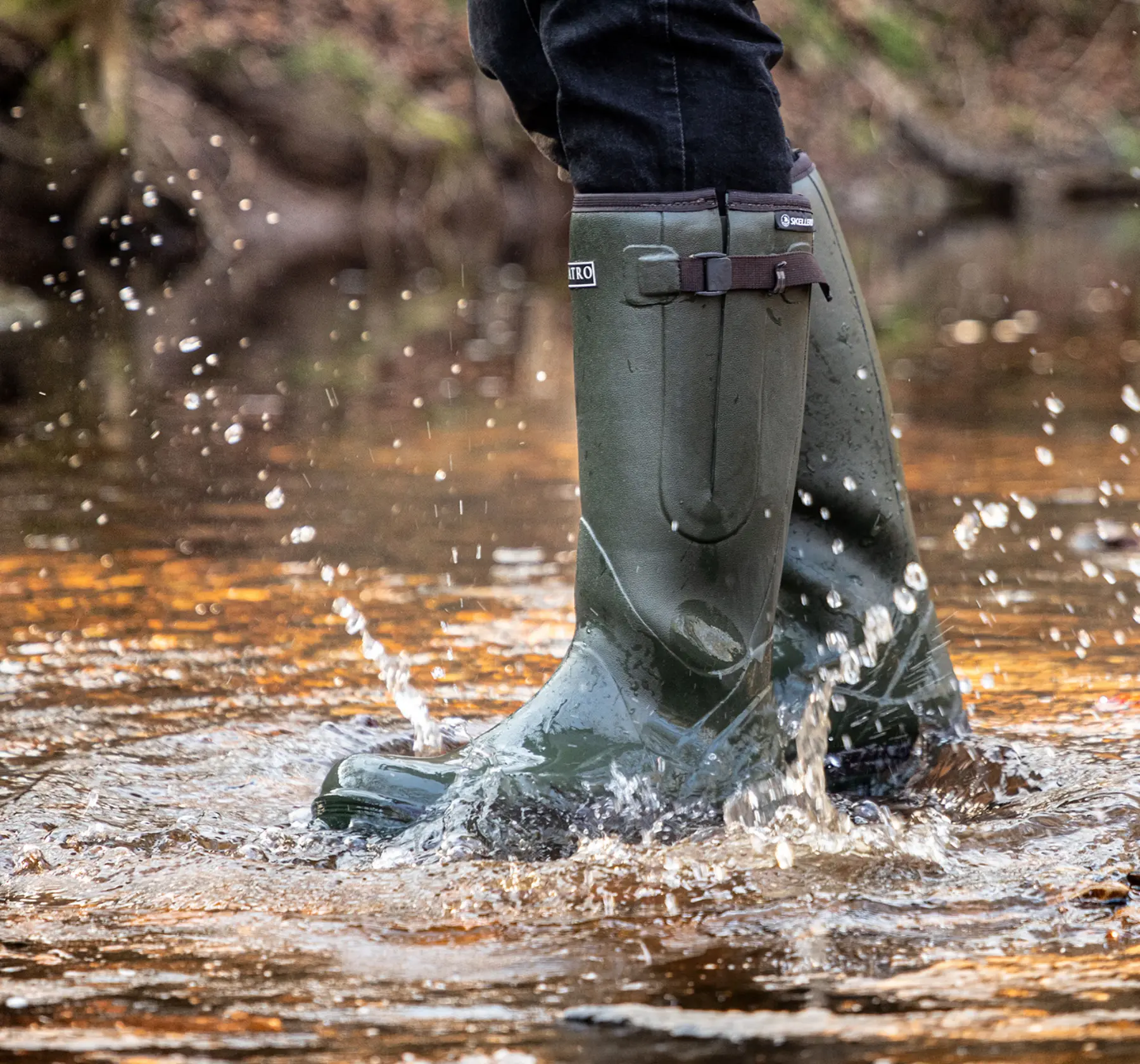 Person wearing tall green rubber boots splashes through shallow water, causing droplets to fly. The background is blurred, showing an outdoor natural setting.