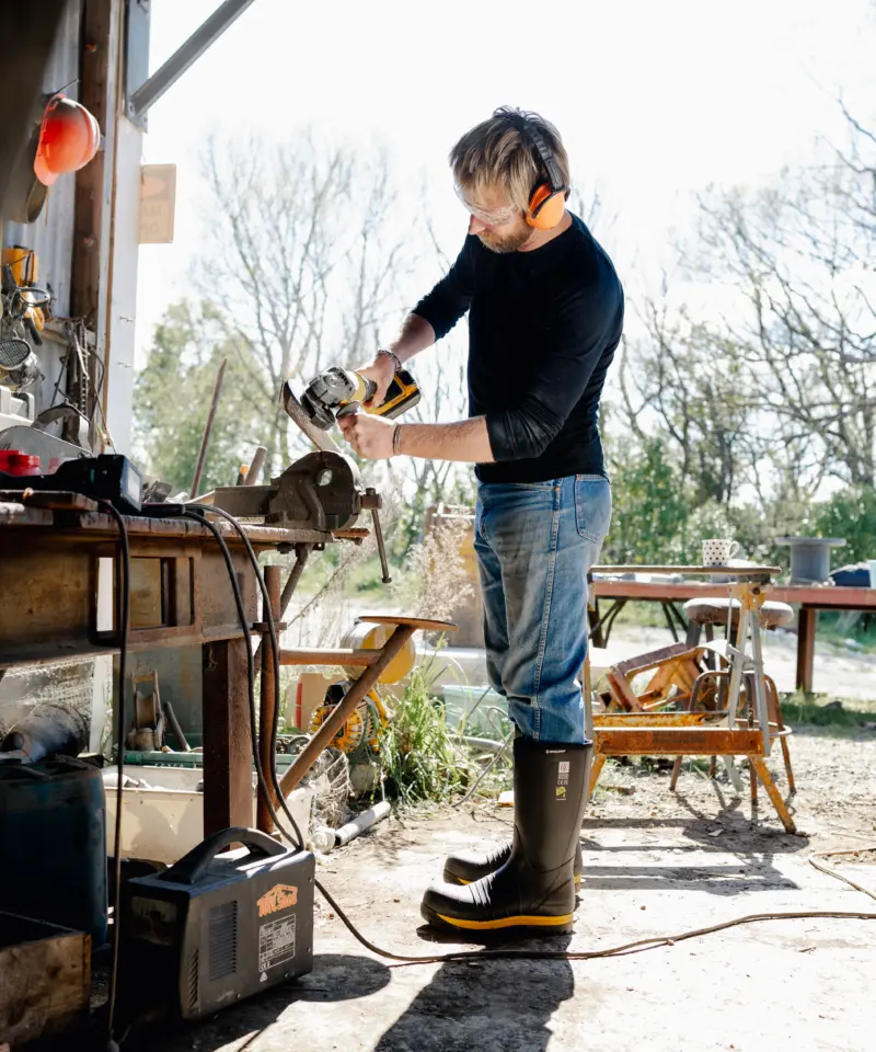 A person wearing rubber boots uses a power tool at a workbench inside a sunny, open workshop with tools and materials scattered around. Trees and greenery are visible outside through the open door.