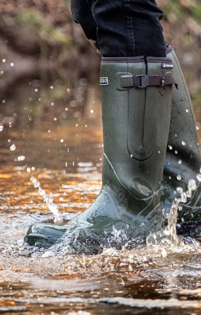 Person wearing tall green rubber boots splashes through shallow water, causing droplets to fly. The background is blurred, showing an outdoor natural setting.