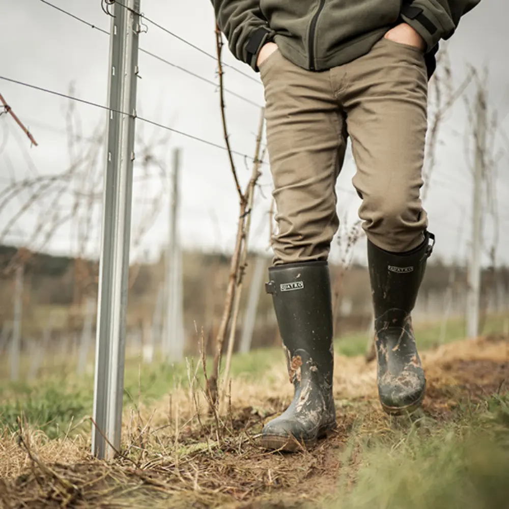 A person wearing muddy black rubber boots and khaki pants walks through a vineyard, with rows of bare grapevines and metal posts visible in the background.