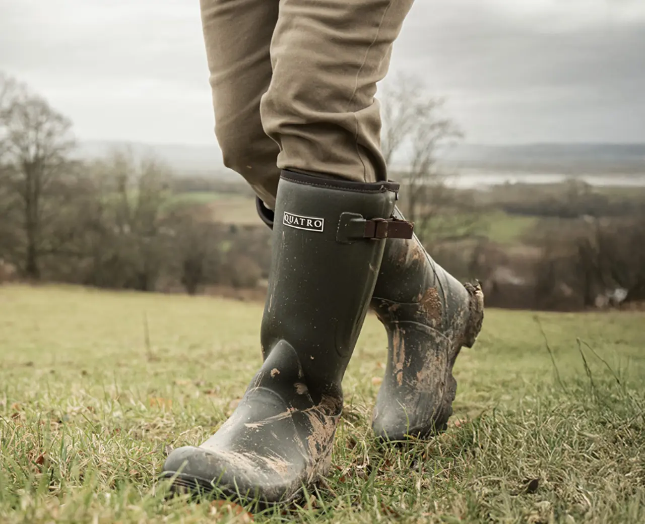 A person wearing muddy green Quatro boots and khaki pants walks on grassy terrain with a scenic, cloudy landscape in the background.