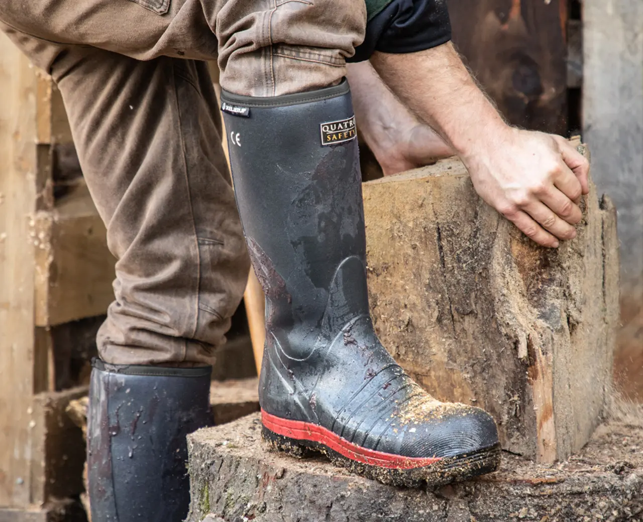 A person wearing muddy rubber boots and brown pants is splitting a large log outdoors, using both hands to position the wood on a tree stump. Wood chips and stacked firewood are visible around them.
