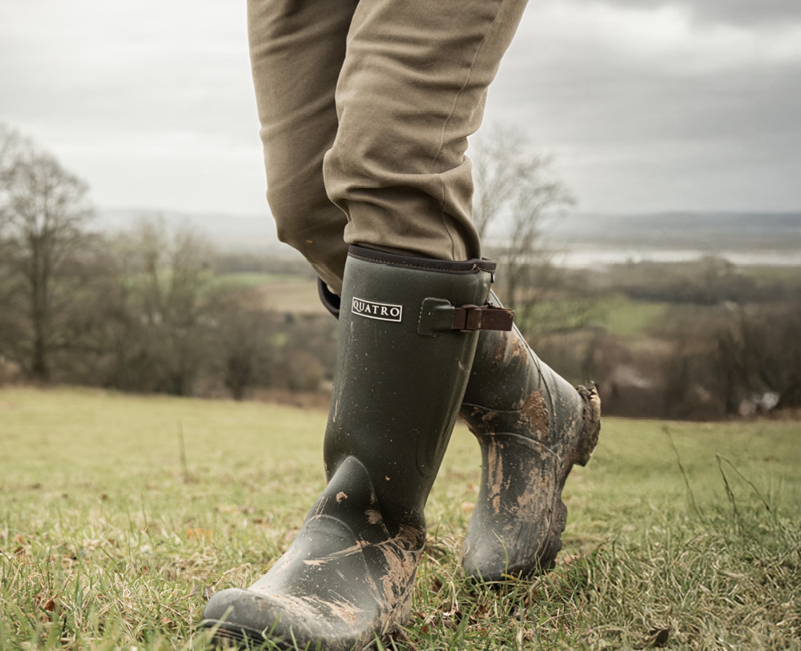 A person wearing muddy green Quatro boots and khaki pants walks on grassy terrain with a scenic, cloudy landscape in the background.