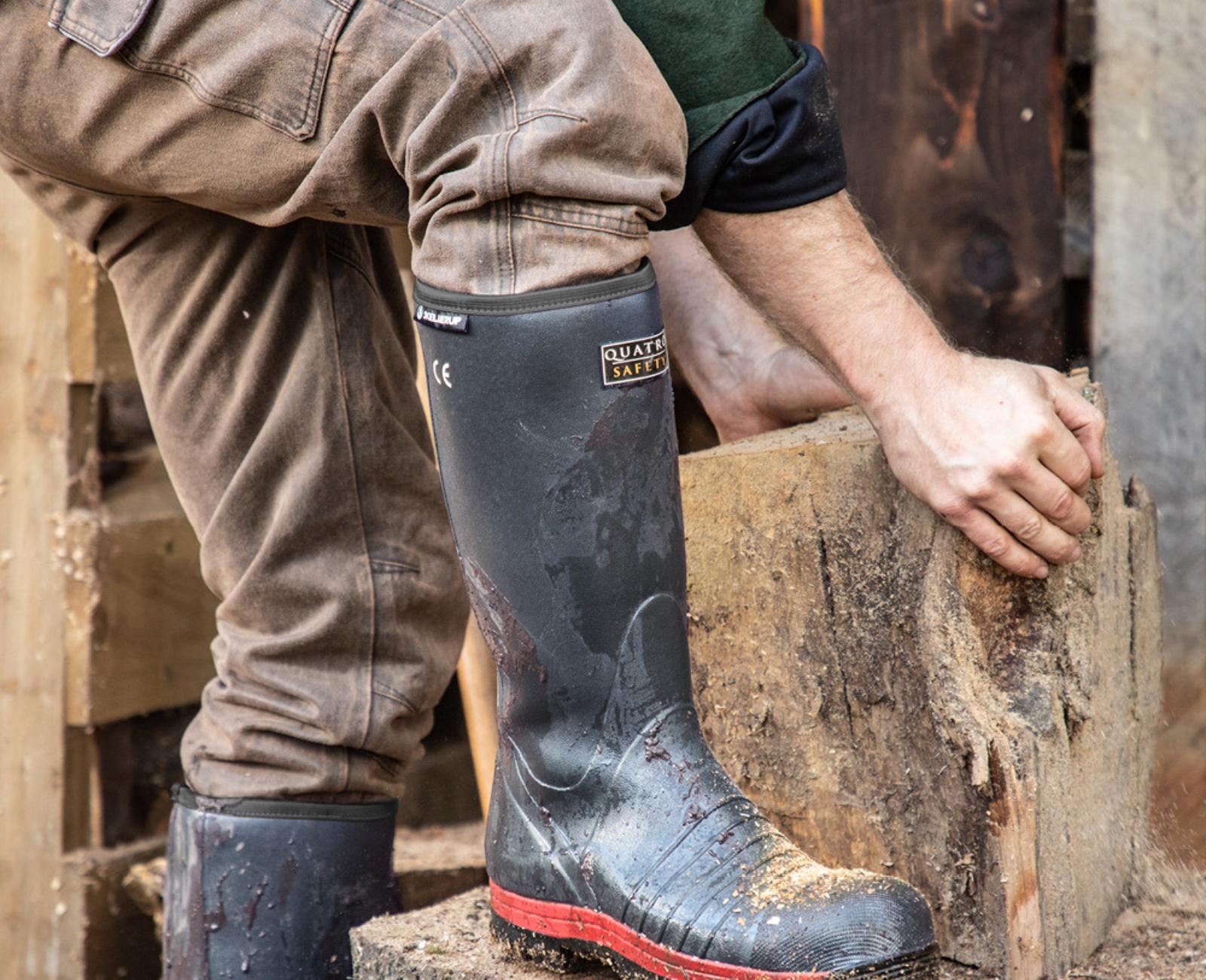A person wearing muddy rubber boots and brown pants is splitting a large log outdoors, using both hands to position the wood on a tree stump. Wood chips and stacked firewood are visible around them.