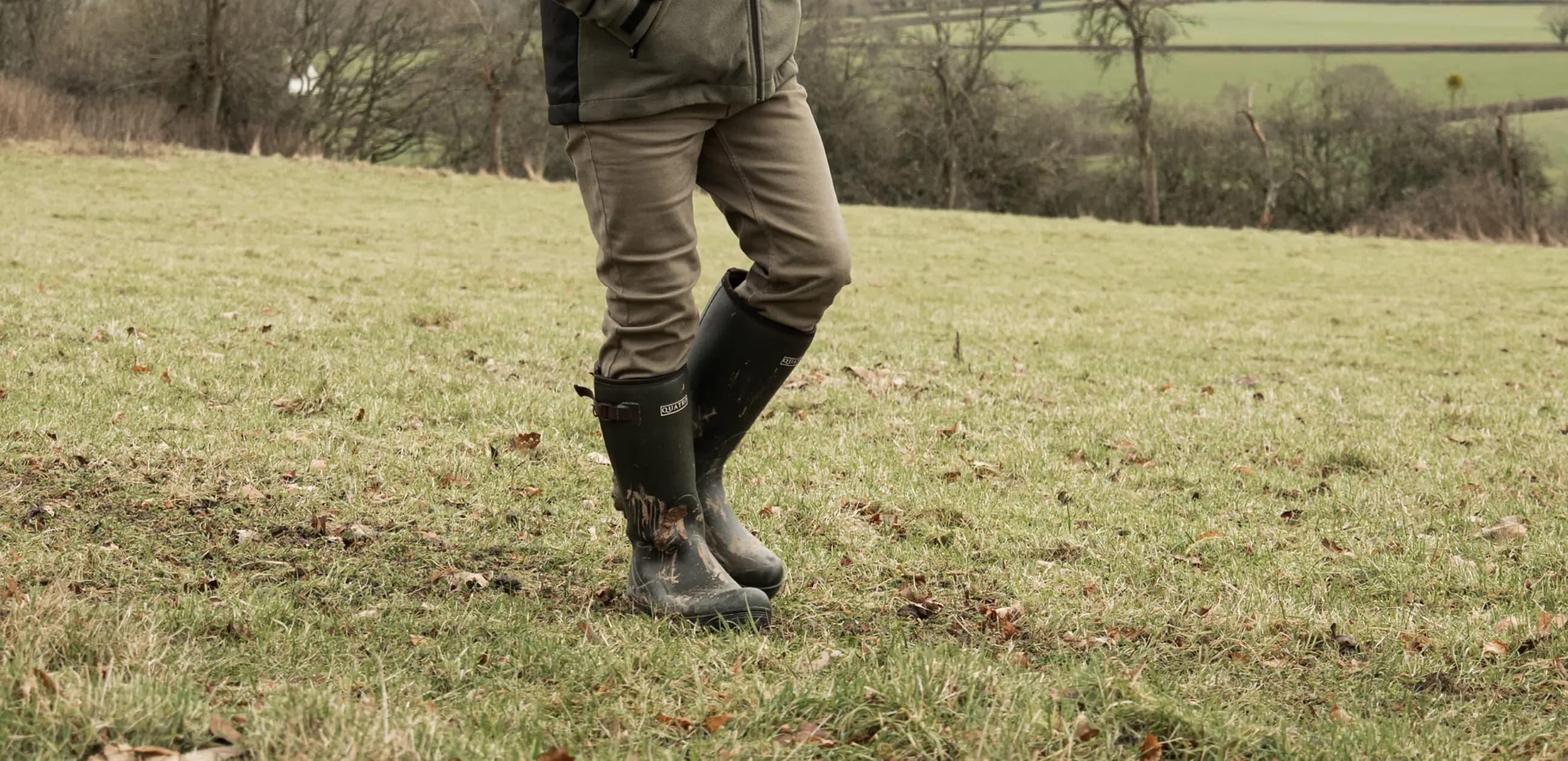 A person standing in a grassy field wearing muddy black boots, tan trousers, and a green jacket. The background shows fields, trees, and hedges under an overcast sky. The persons upper body is out of frame.