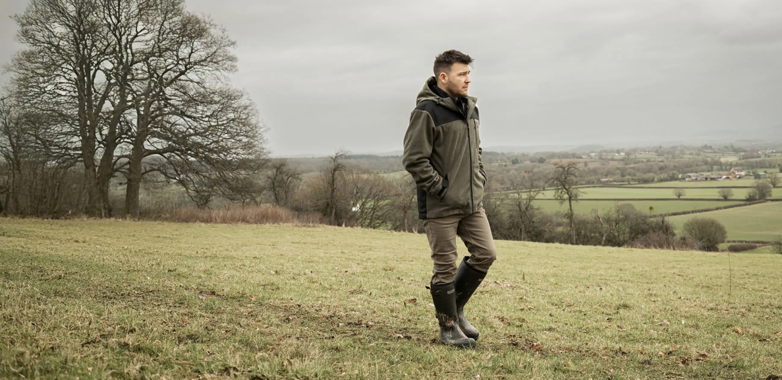 A man in a green jacket and boots stands in a grassy field on a cloudy day, with bare trees and rolling countryside visible in the background.