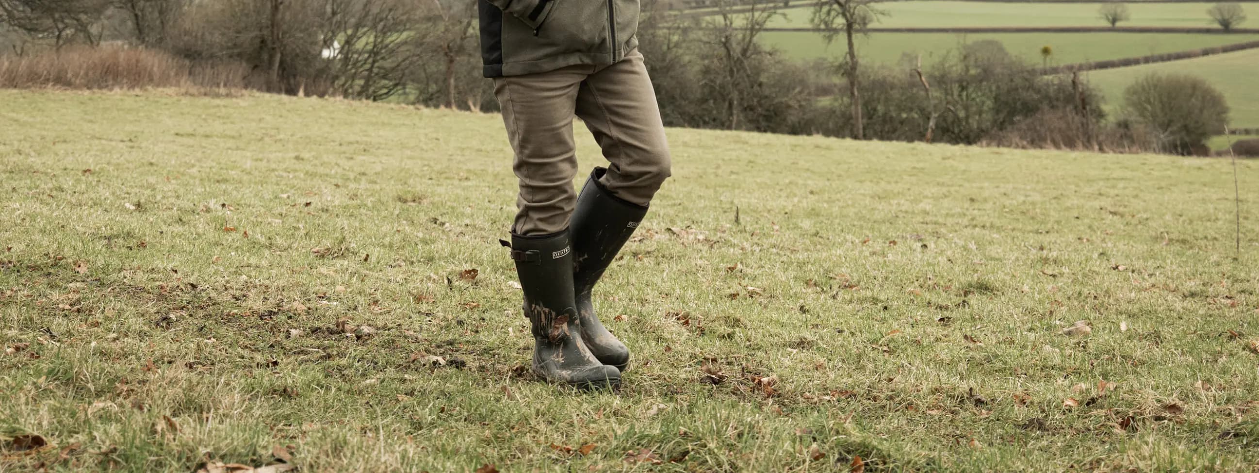 A person standing in a grassy field wearing muddy black boots, tan trousers, and a green jacket. The background shows fields, trees, and hedges under an overcast sky. The persons upper body is out of frame.