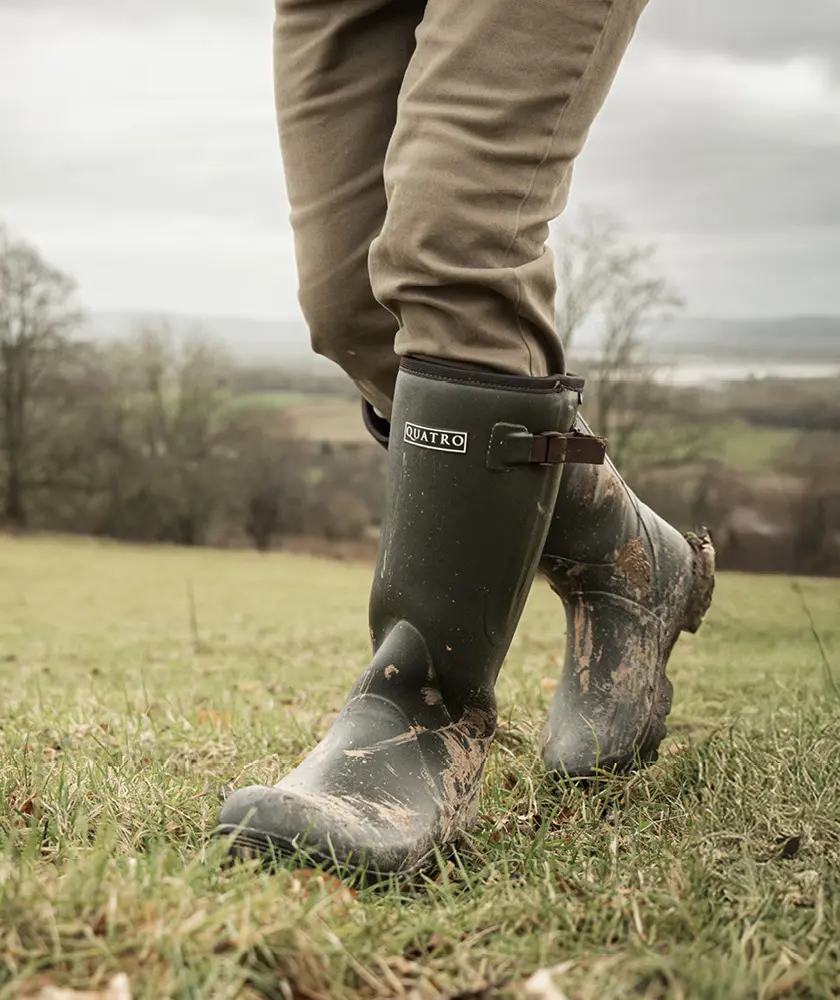 A person wearing muddy green Quatro boots and khaki pants walks on grassy terrain with a scenic, cloudy landscape in the background.