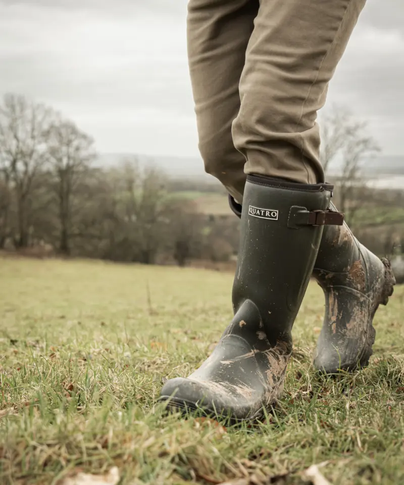 A person wearing muddy rubber boots and khaki pants walks through a grassy, open field on a cloudy day, with bare trees and distant hills in the background.