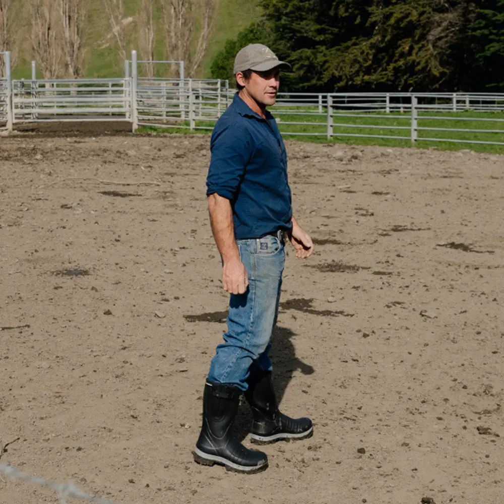 A man wearing a cap, blue shirt, jeans, and black rubber boots stands on dirt in a fenced outdoor area, possibly a farm or ranch, with trees and grass in the background.