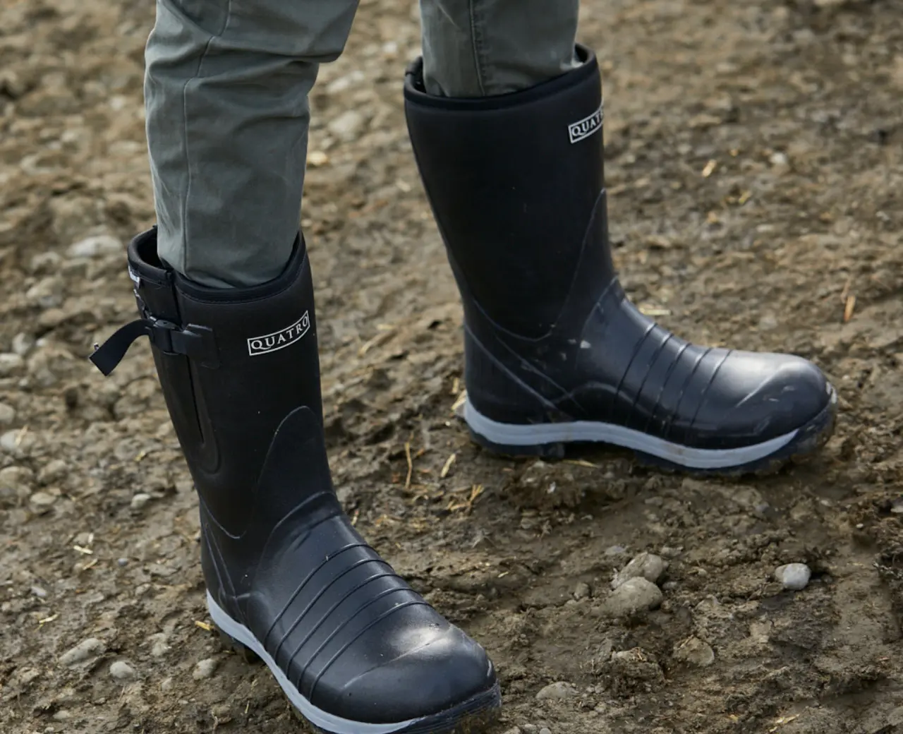 Person wearing black rubber boots and green pants standing on uneven, muddy ground.