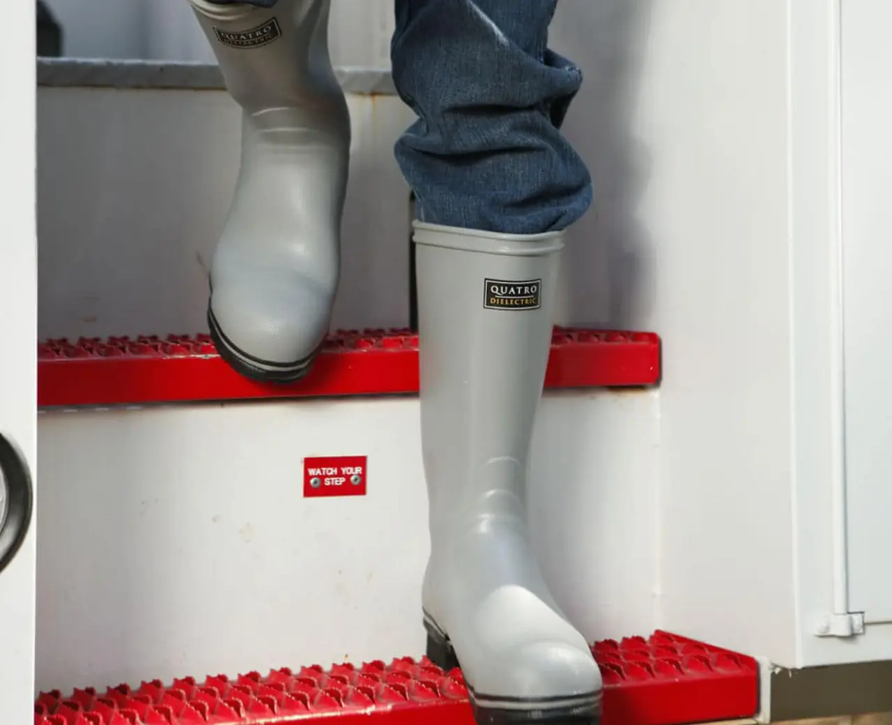 A person wearing gray rubber safety boots and blue jeans steps down red metal stairs from a white vehicle or structure.