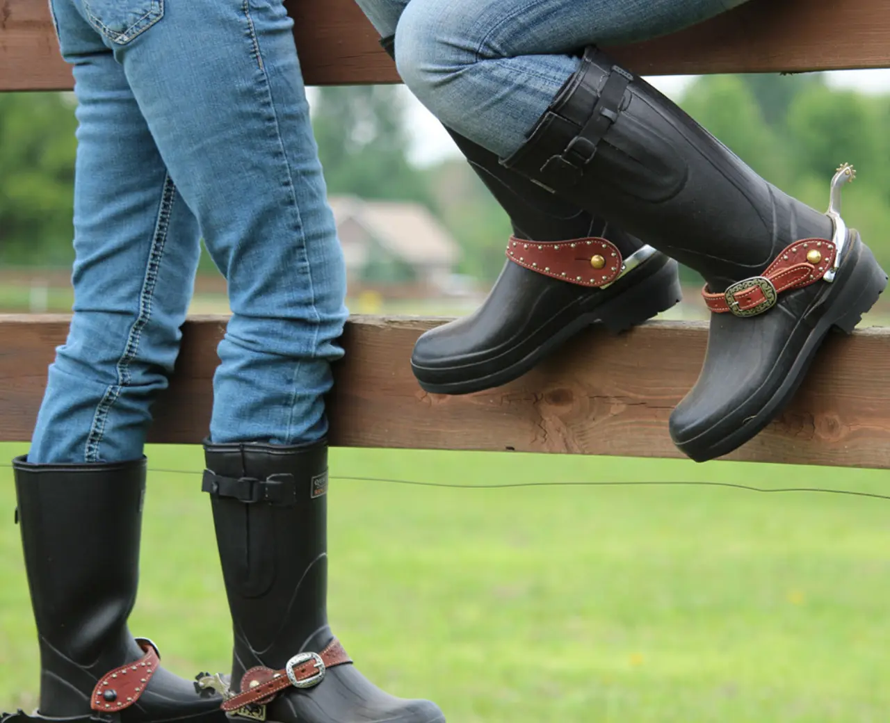 Two people wearing blue jeans, black rubber boots, and brown leather spurs sit on a wooden fence, with green grass and trees visible in the background.