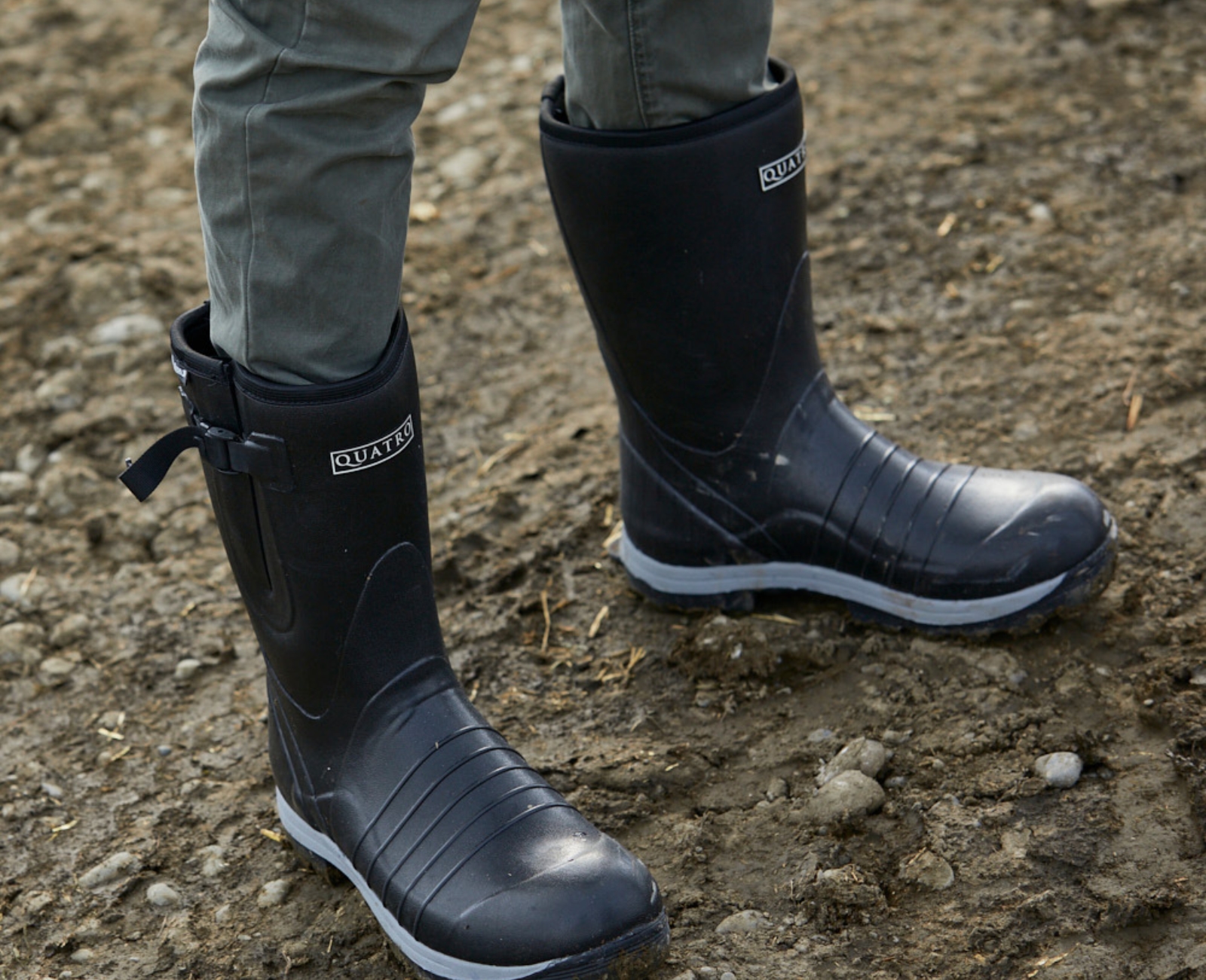Person wearing black rubber boots and green pants standing on uneven, muddy ground.