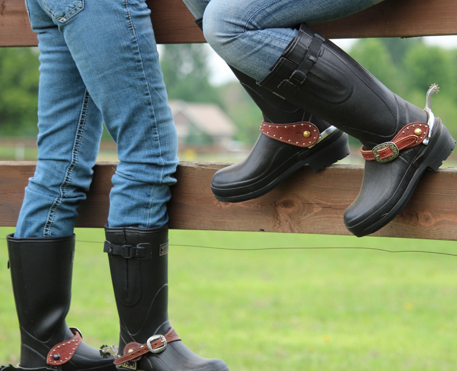 Two people wearing blue jeans, black rubber boots, and brown leather spurs sit on a wooden fence, with green grass and trees visible in the background.