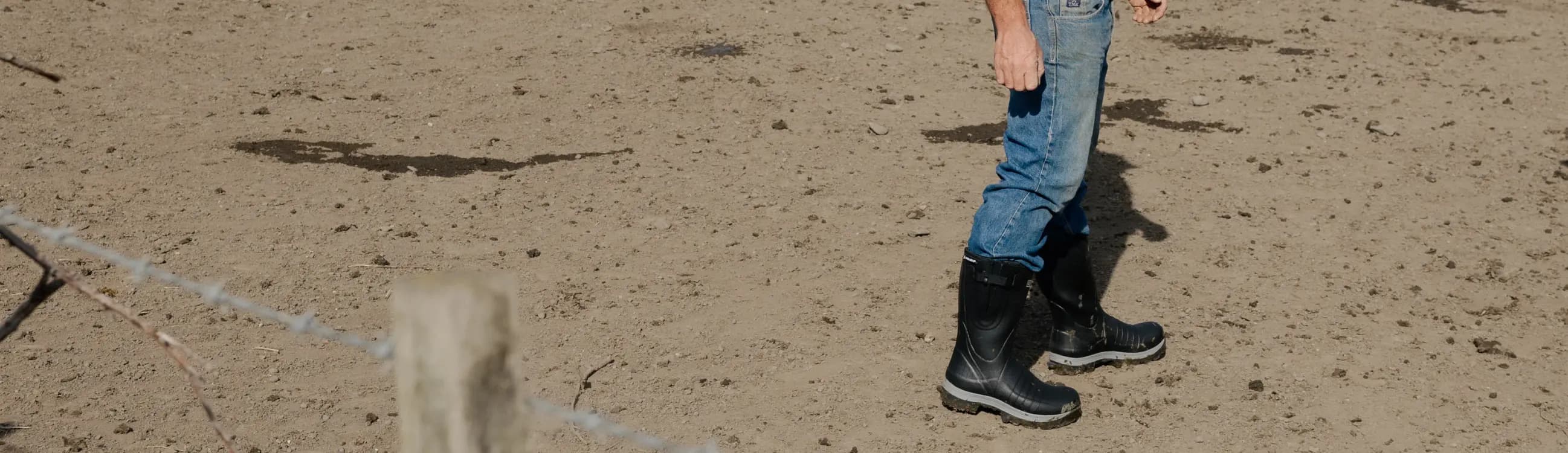 A person wearing blue jeans and black rubber boots stands on dry, sandy ground near a barbed wire fence post. Only the lower half of their body is visible.