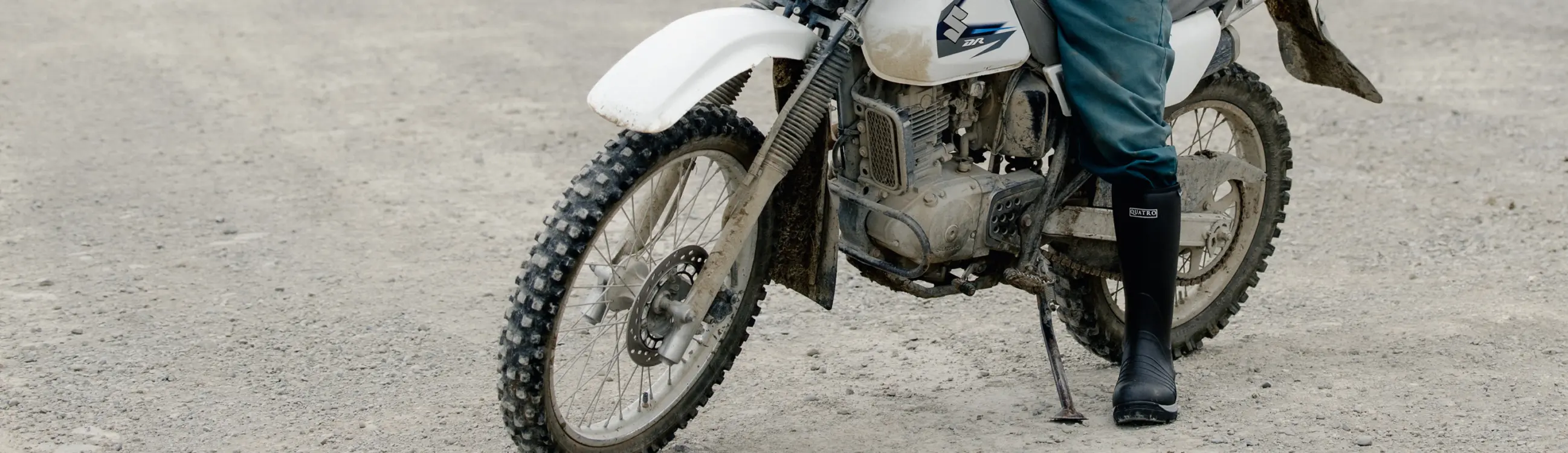 A person wearing black boots and teal pants stands next to a white dirt bike on a gravel surface, with only their lower body and the motorcycle visible.