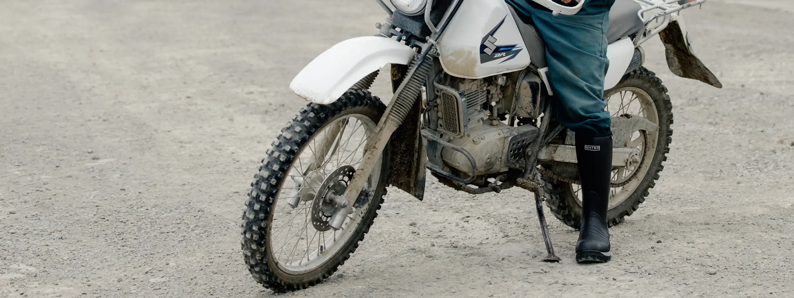 A person wearing black boots and teal pants stands next to a white dirt bike on a gravel surface, with only their lower body and the motorcycle visible.
