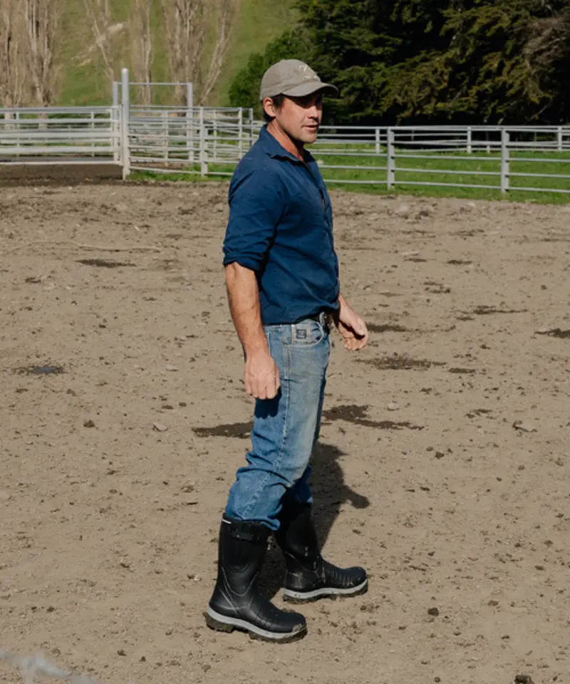 A man wearing a cap, blue shirt, jeans, and black rubber boots stands on dirt in a fenced outdoor area, possibly a farm or ranch, with trees and grass in the background.