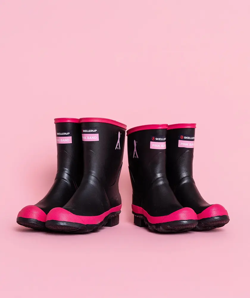 Two pairs of black rain boots with pink soles and trim are placed side by side on a pink background. Each boot is decorated with a pink ribbon symbol, indicating support for breast cancer awareness.