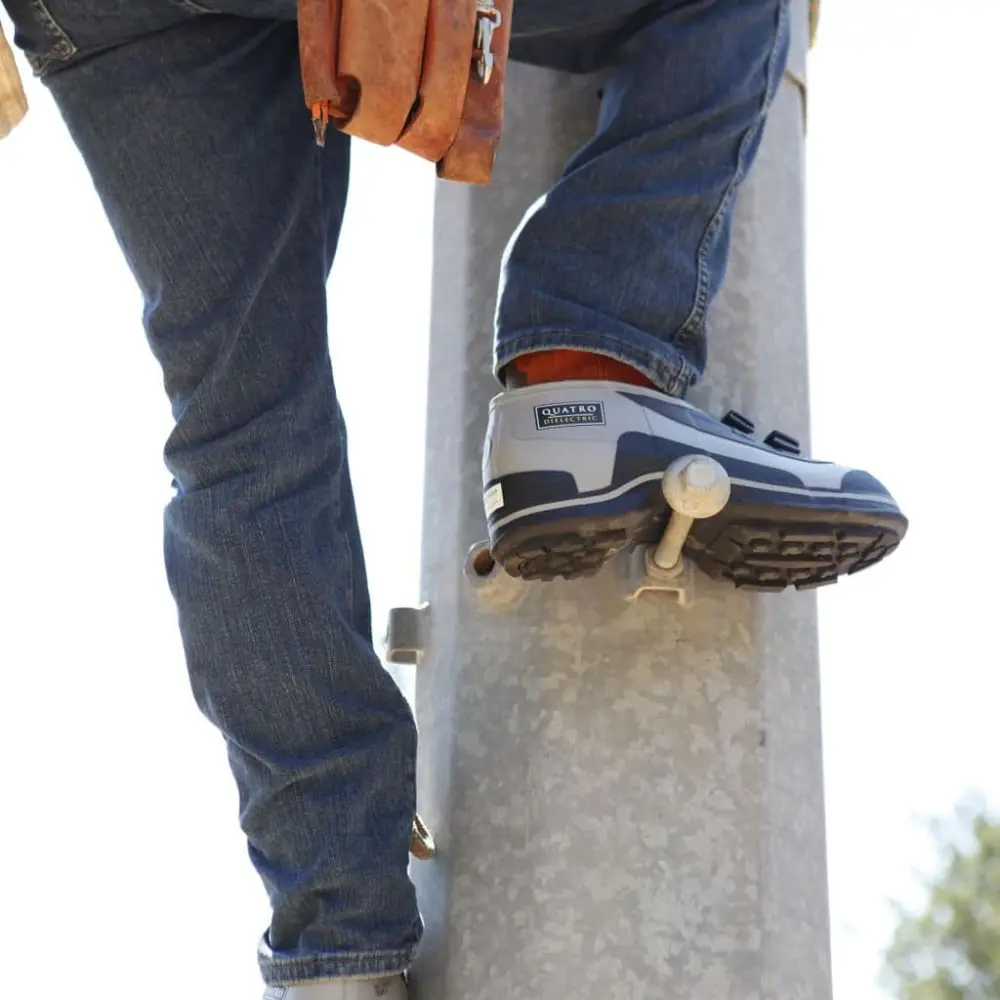 A utility worker wearing jeans, safety shoes, gloves, and a tool belt climbs a concrete pole using metal climbing spikes attached to the pole. The image focuses on the workers lower body and climbing equipment.