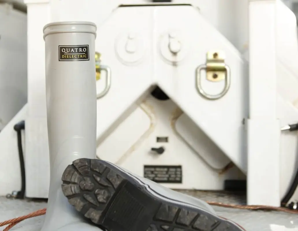 A pair of gray QUATRO protective rubber boots, with one boot upright and the other lying on its side, are placed on a metallic platform in front of industrial equipment and a coil of orange cable.