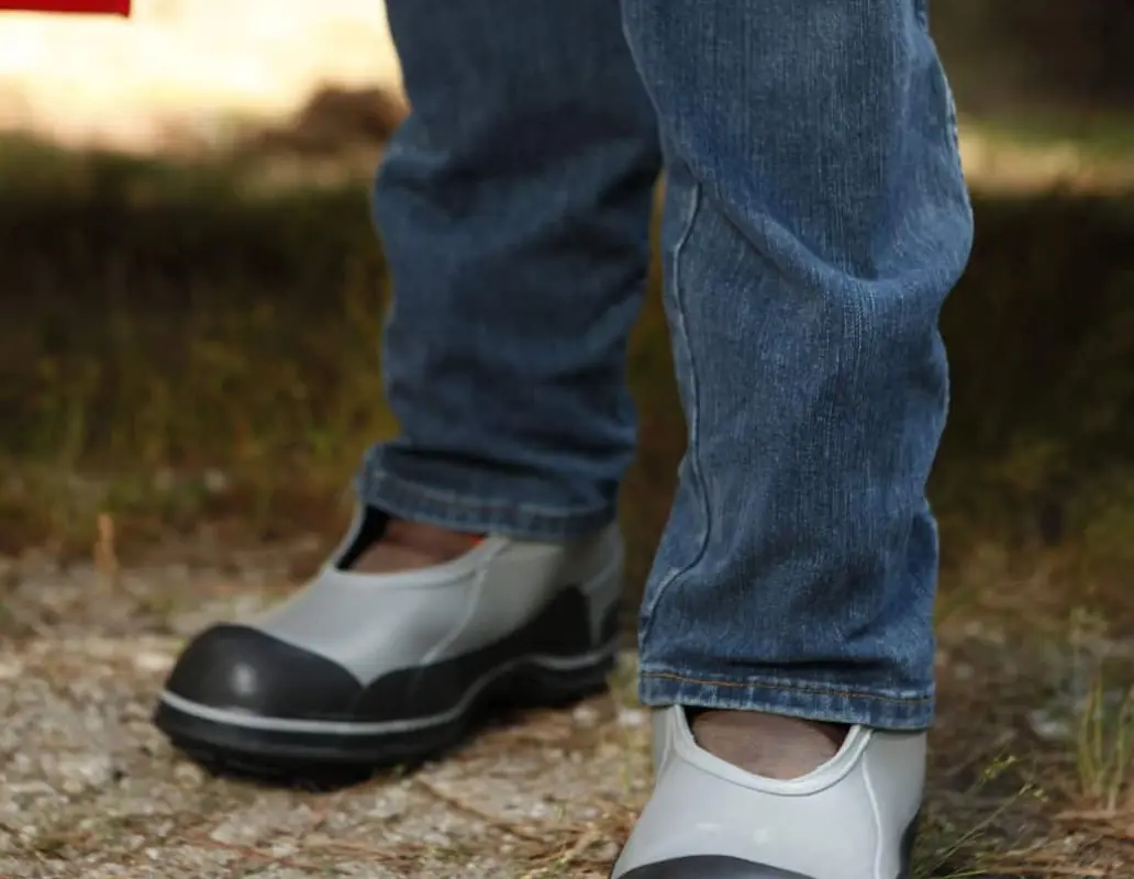 A person wearing blue jeans and gray slip-on shoes stands on a dirt and grass surface outdoors. Only the legs and feet are visible.