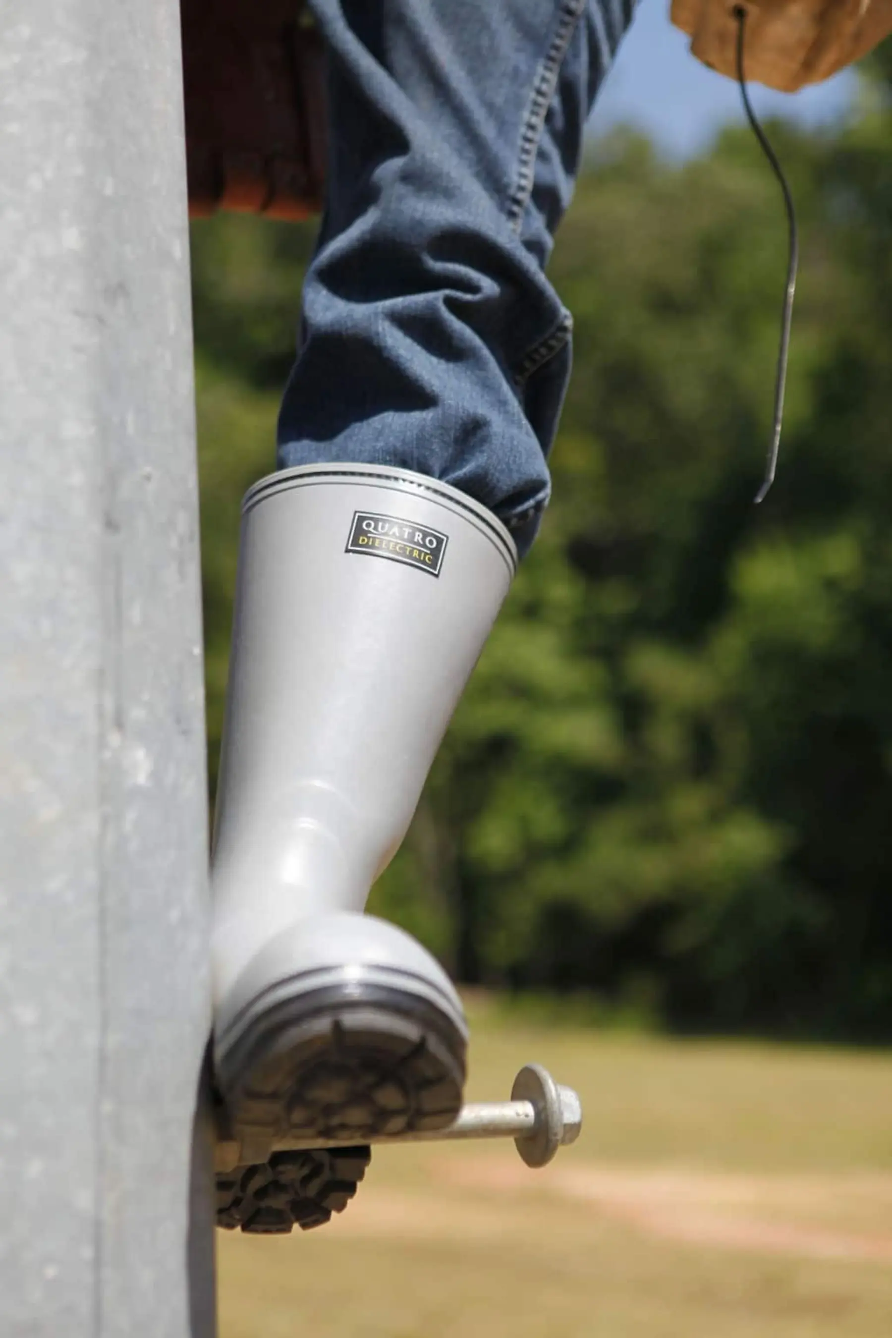 A person wearing blue jeans and light gray rubber boots steps onto a metal surface outdoors, with trees and grass visible in the background.
