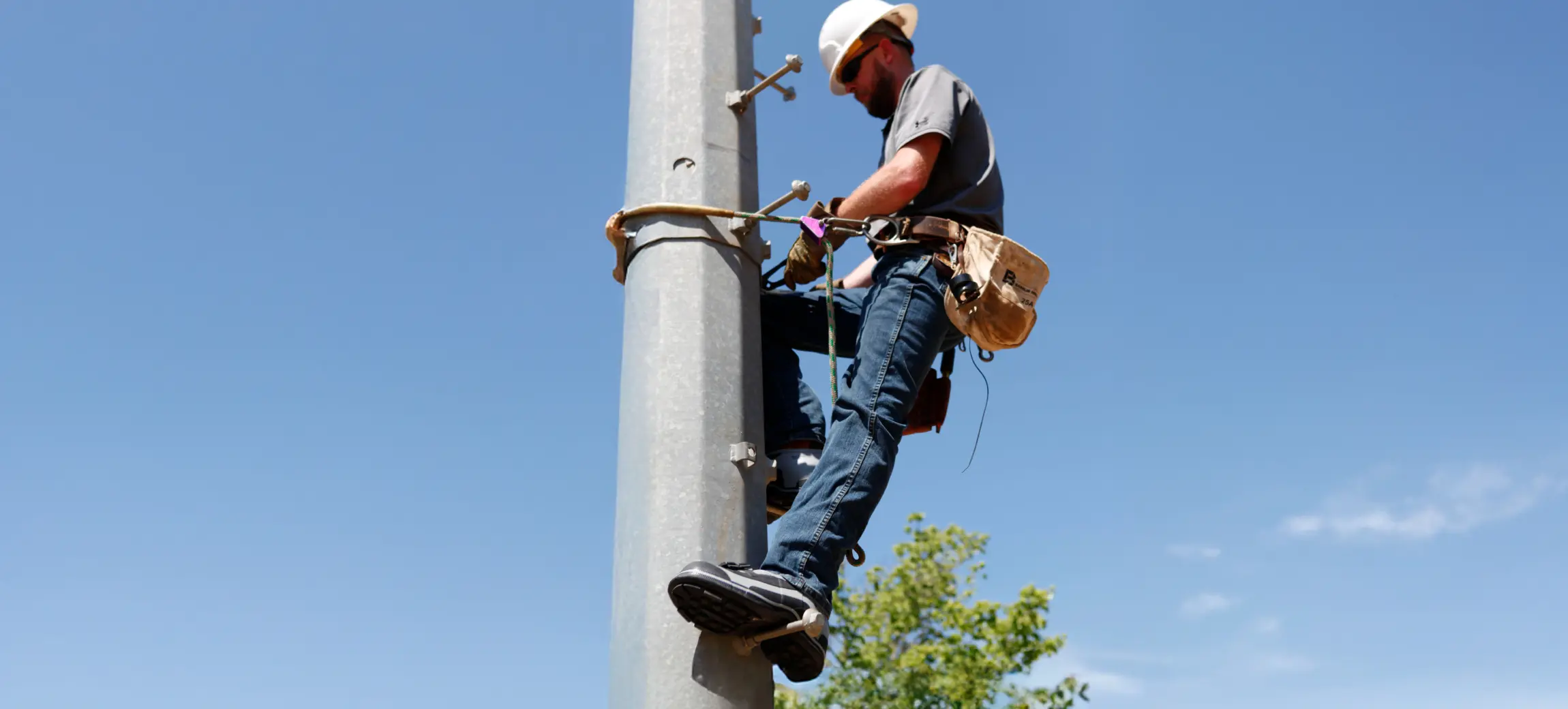 A worker wearing a hard hat and safety harness climbs a utility pole against a clear blue sky, carrying tools in a belt around his waist.