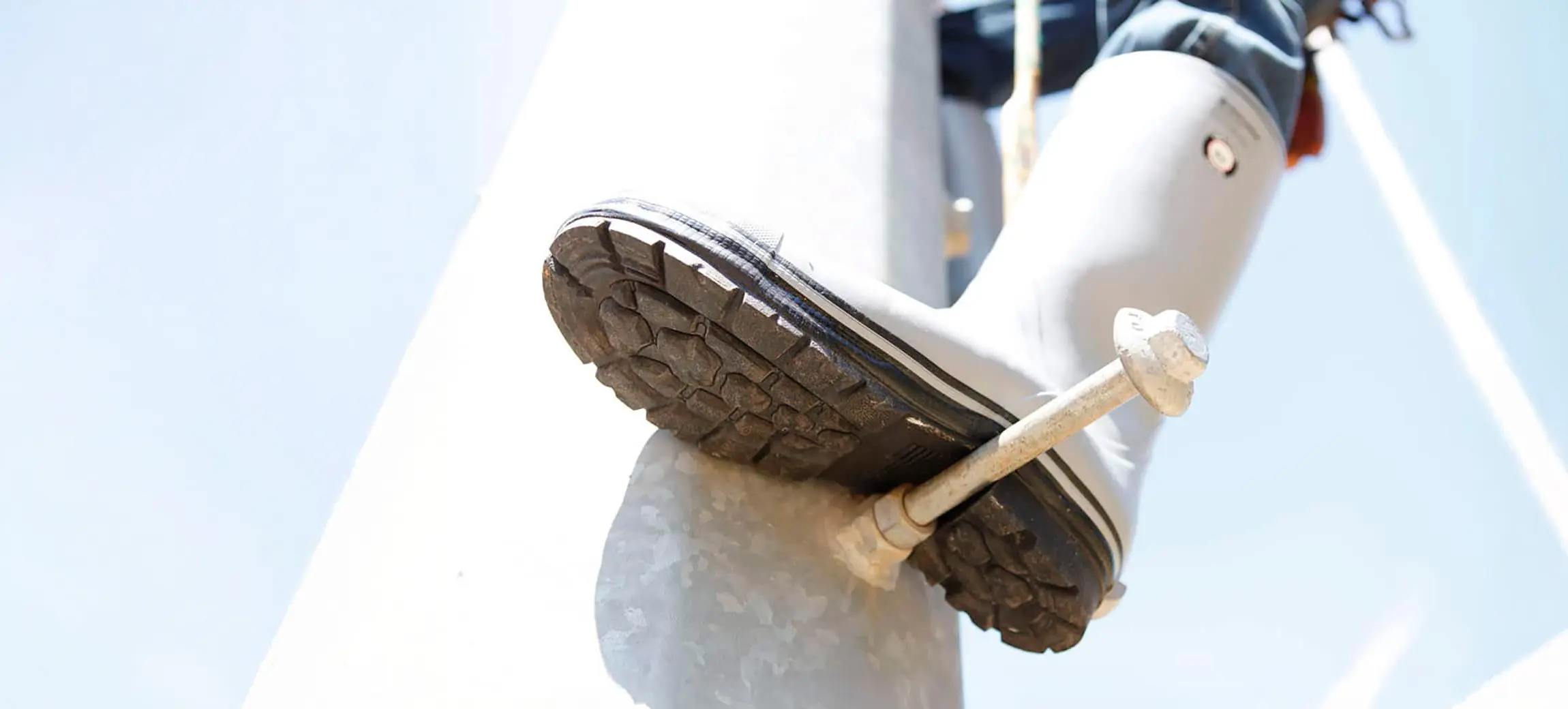 Close-up of a person wearing a rubber boot, stepping on a metal pole with a metal step attached, viewed from below against a bright sky.