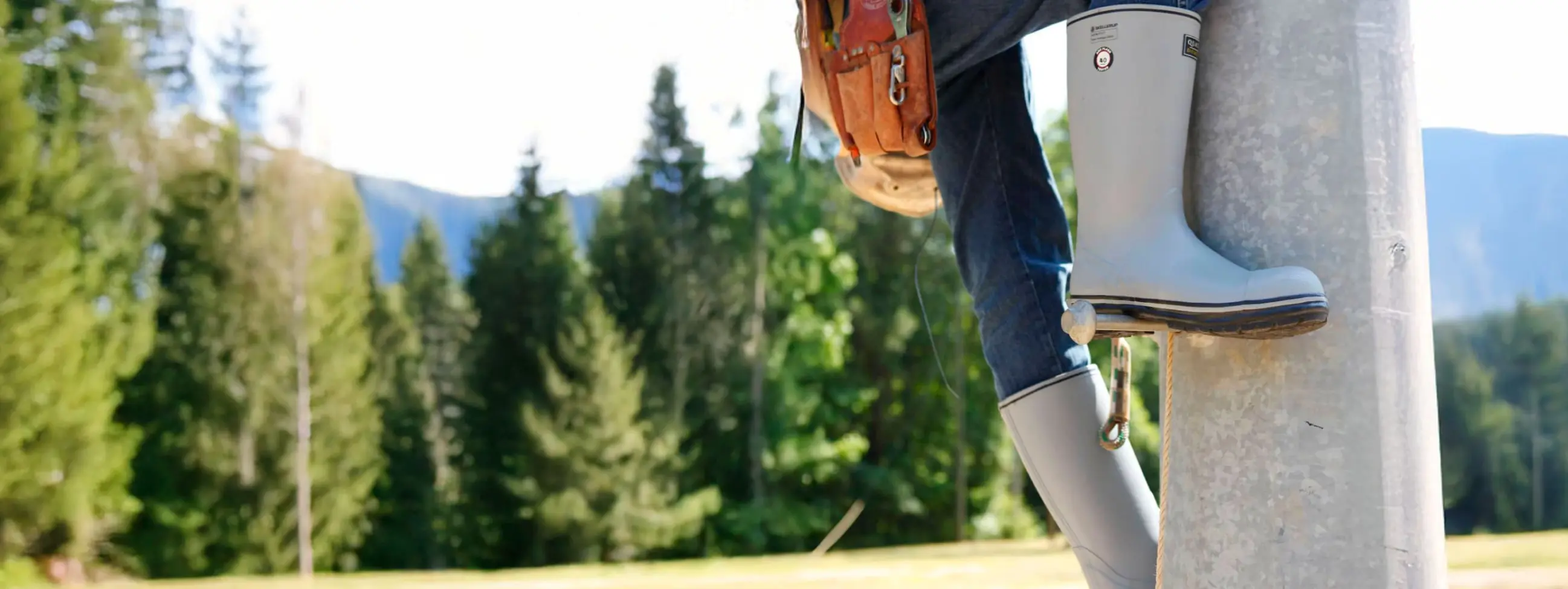 A person wearing a tool belt, jeans, and gray work boots climbs a utility pole with climbing spikes. Pine trees and mountains are visible in the background under a clear sky.