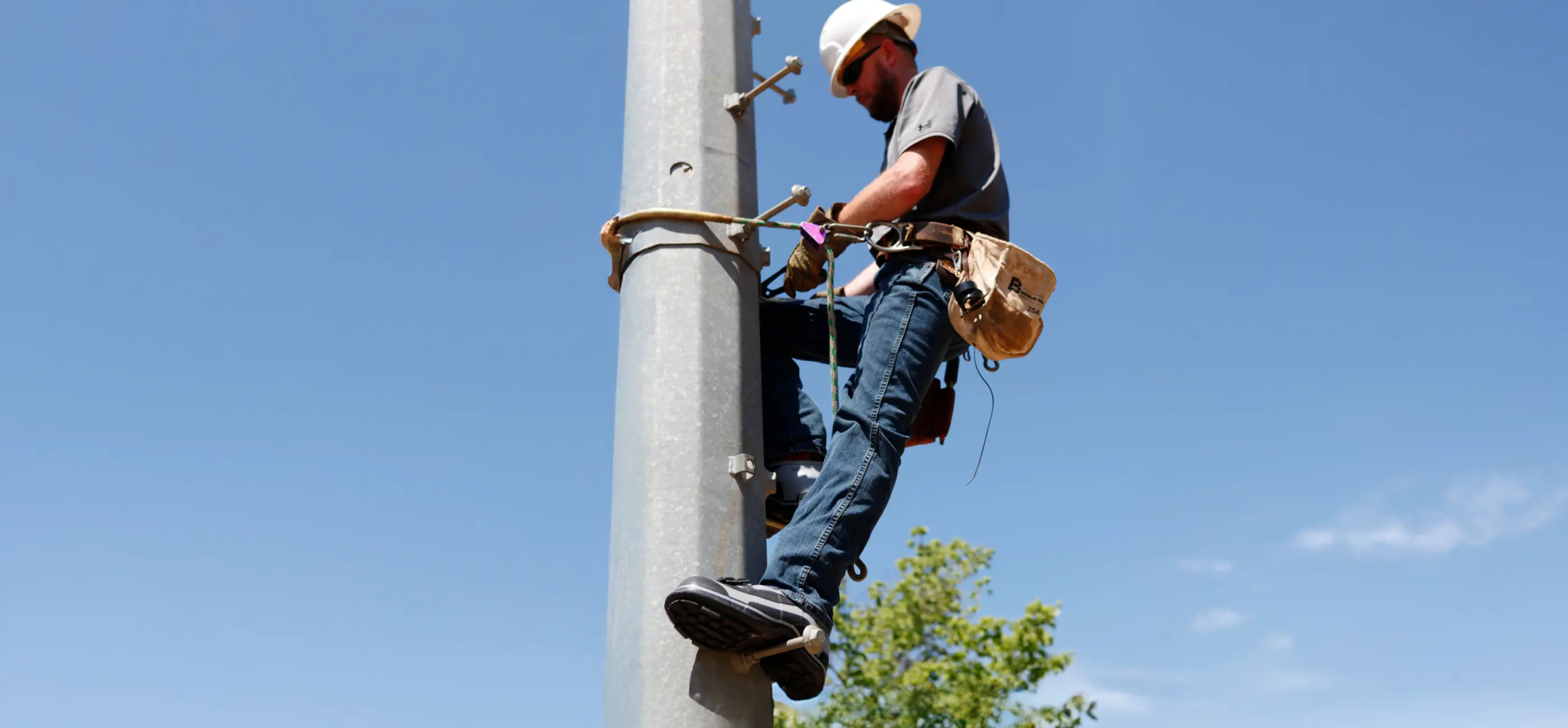 A worker wearing a hard hat and safety harness climbs a utility pole against a clear blue sky, carrying tools in a belt around his waist.