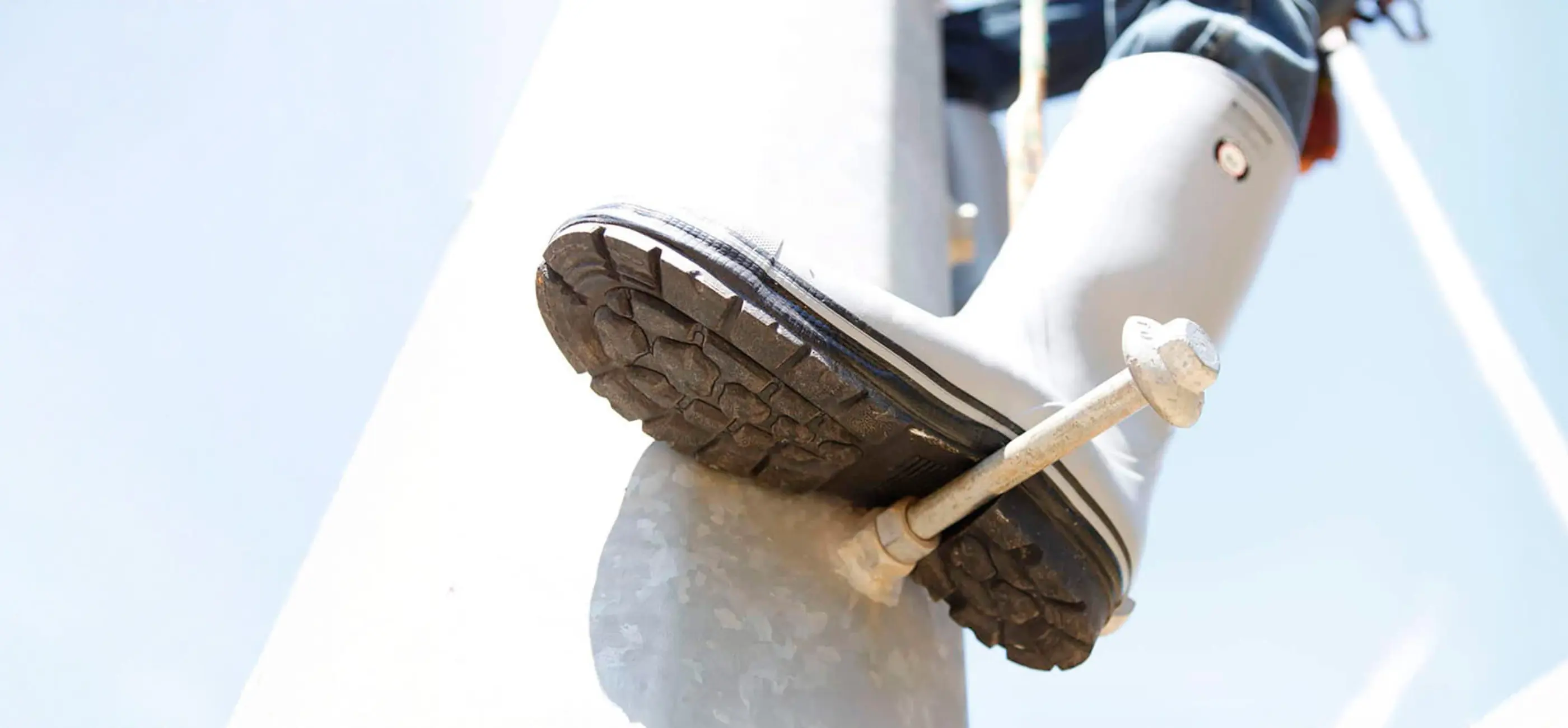Close-up of a person wearing a rubber boot, stepping on a metal pole with a metal step attached, viewed from below against a bright sky.