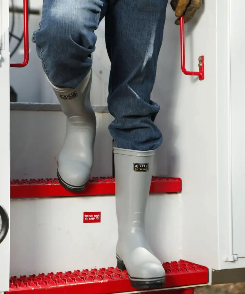 A person wearing gray rubber boots, blue jeans, and a glove steps down red metal stairs from a white vehicle or structure, holding onto a red handrail.