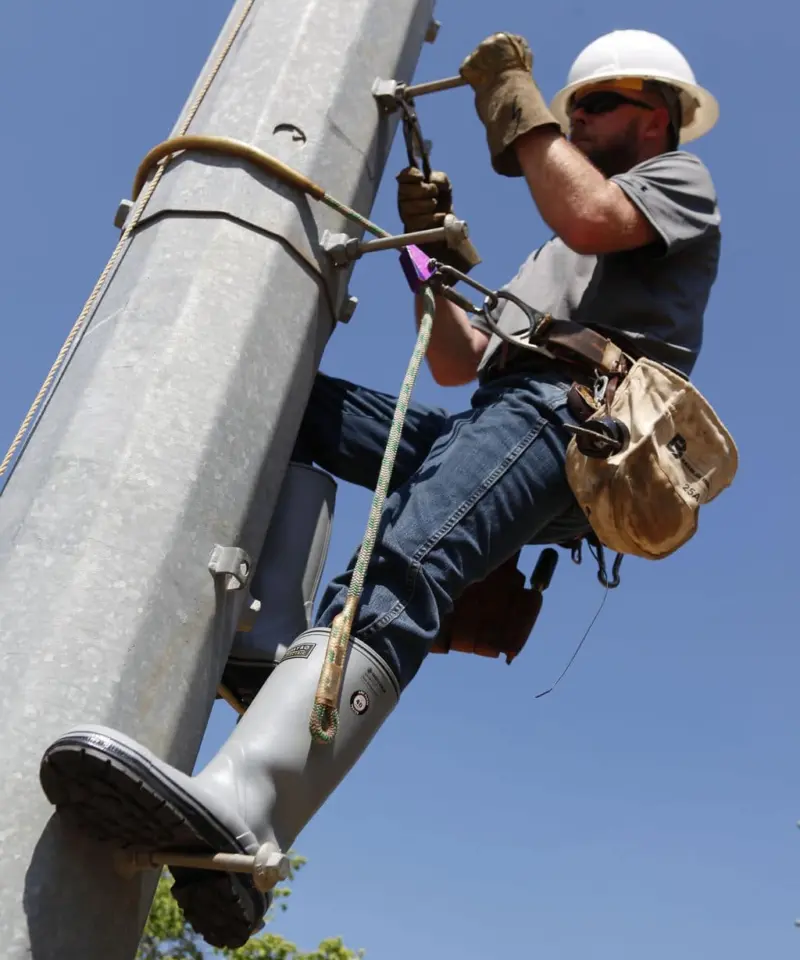 A utility worker wearing a hard hat, gloves, boots, and safety gear climbs a utility pole with tools, working under a clear blue sky.
