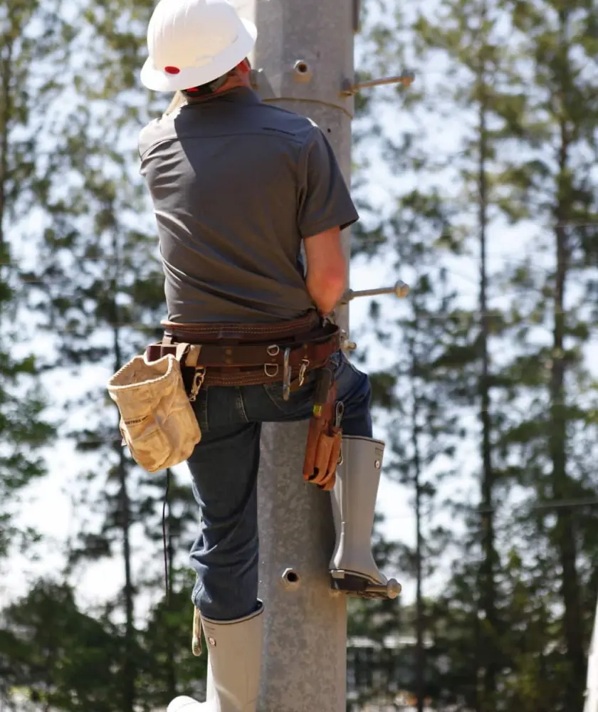 A utility worker wearing a hard hat and tool belt climbs a utility pole outdoors, with trees and a blue sky in the background.