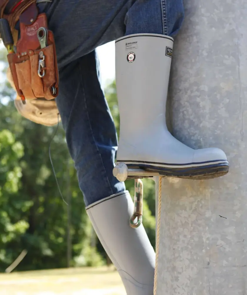A worker wearing blue jeans, gray rubber boots, and a safety harness climbs a utility pole with the aid of climbing equipment on a sunny day.