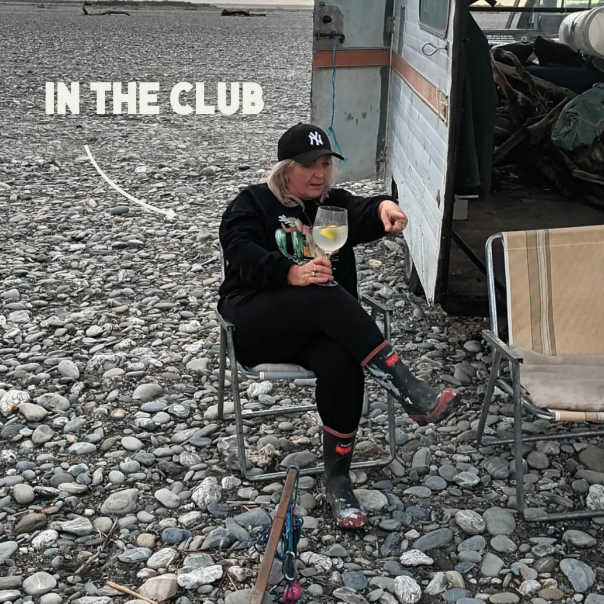 A woman in black clothing and a New York Yankees cap sits on a chair outside, holding a drink, next to a trailer on a rocky ground. The words IN THE CLUB are written on the left side of the image.