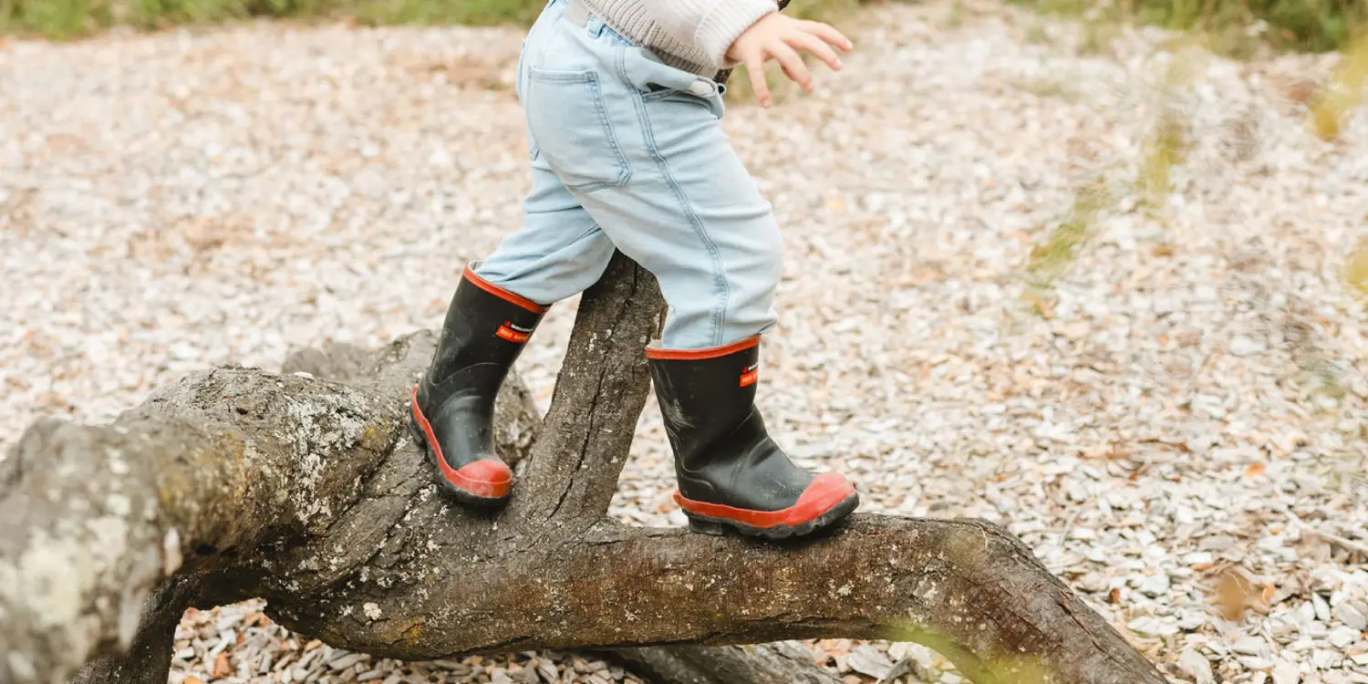 A child in light blue jeans and black rain boots with red soles balances on a fallen tree branch outdoors, surrounded by gravel and leaves.