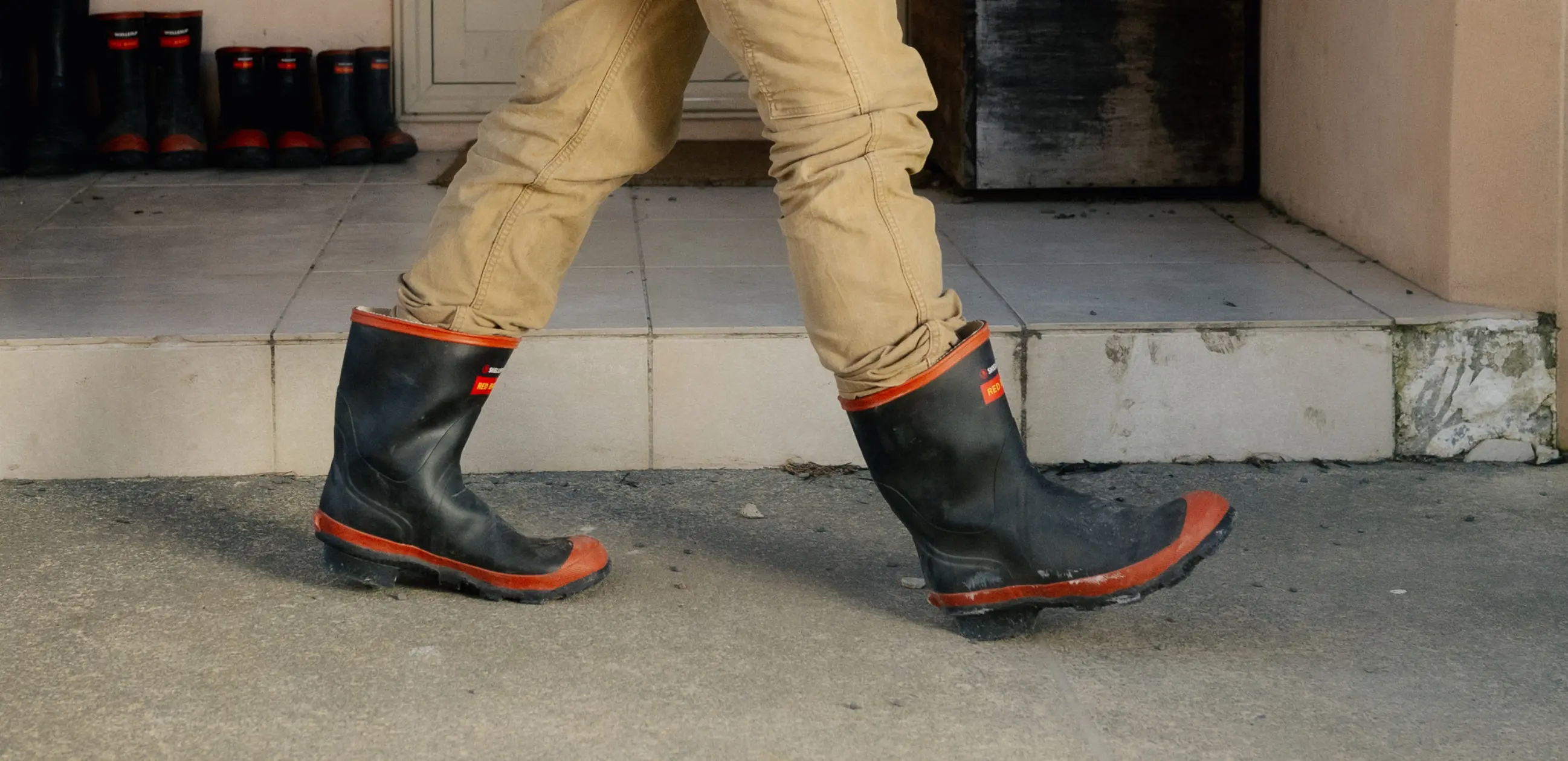 Person wearing tan pants and black rubber boots with red trim walks on a concrete surface outside a building; several pairs of similar boots are lined up on a tiled porch in the background.
