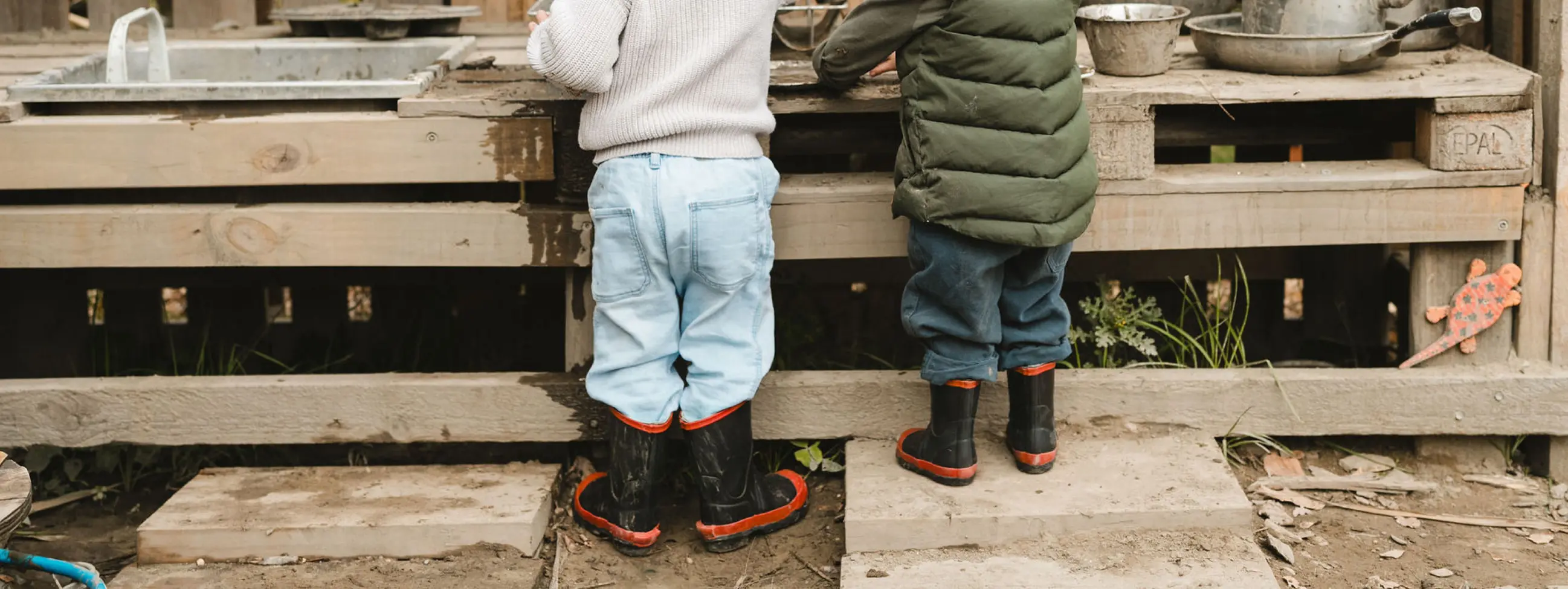Two young children wearing rain boots and outdoor clothes stand at a rustic wooden play kitchen, seen from behind, with metal pots and pans on the counter and dirt and leaves on the ground.