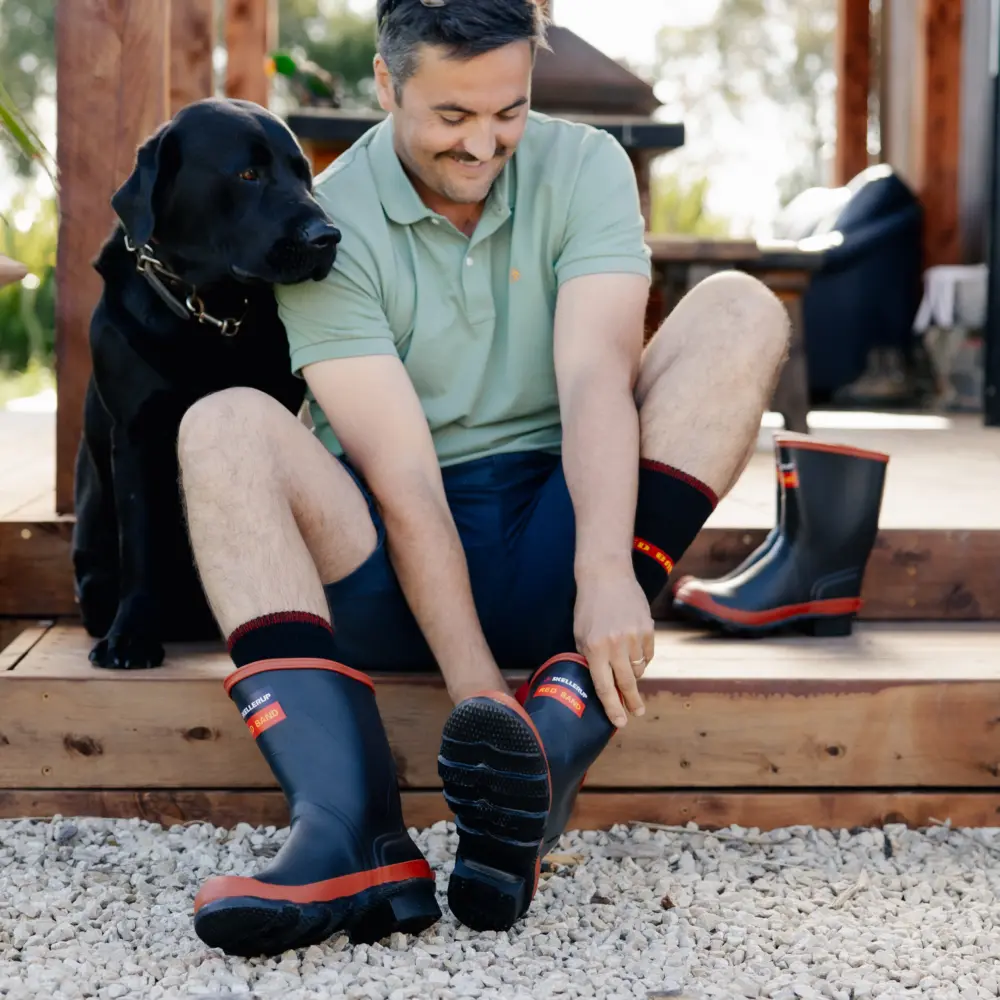 A man sitting on wooden steps outdoors, smiling as he puts on rubber boots. A black dog sits beside him, and another pair of boots is visible in the background on the porch.