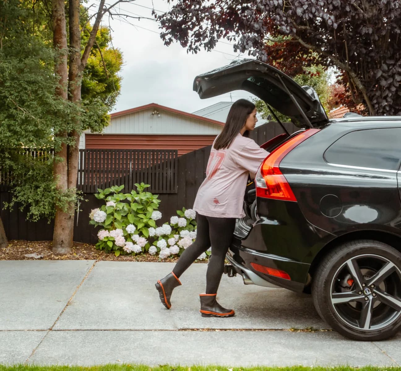 A woman wearing black leggings and rain boots places items into the trunk of a black SUV parked in a driveway, with trees, flowers, and a wooden fence in the background.