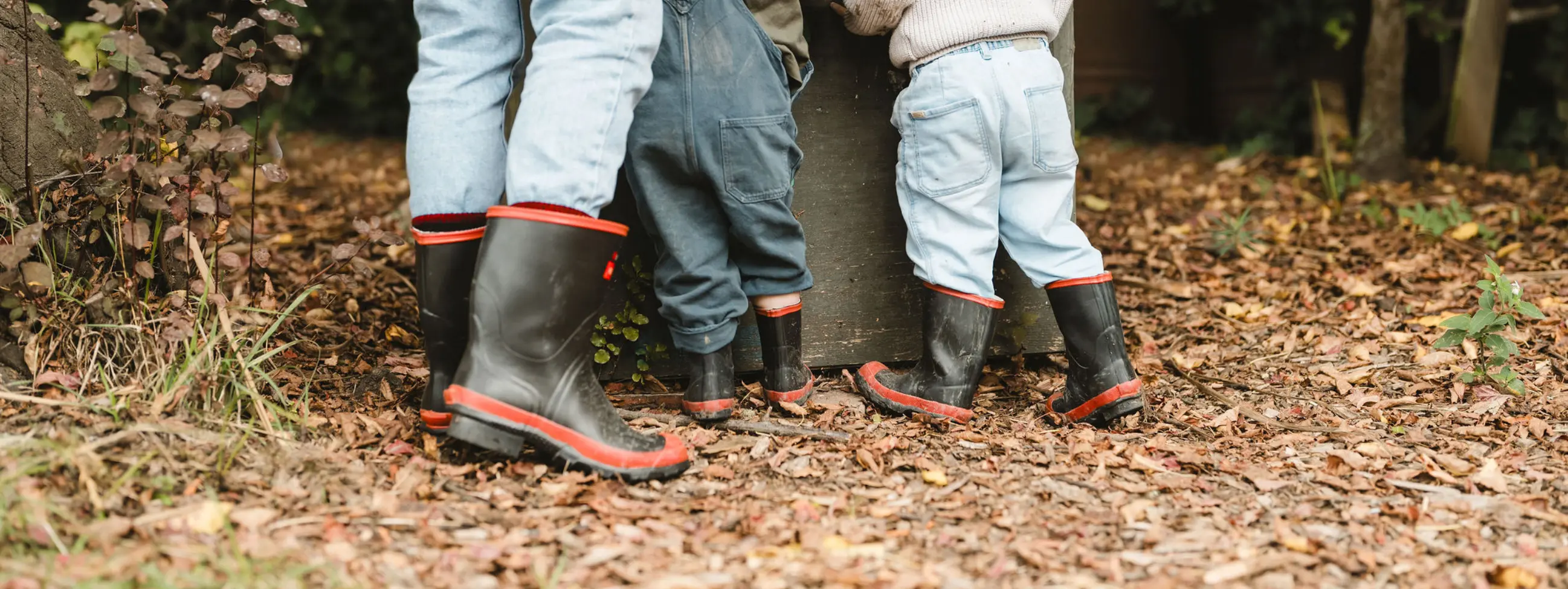 An adult and two young children, all wearing jeans and black rain boots with red trim, stand together outdoors on leafy ground, facing away, as they interact with a large metal container. Trees and plants surround them.
