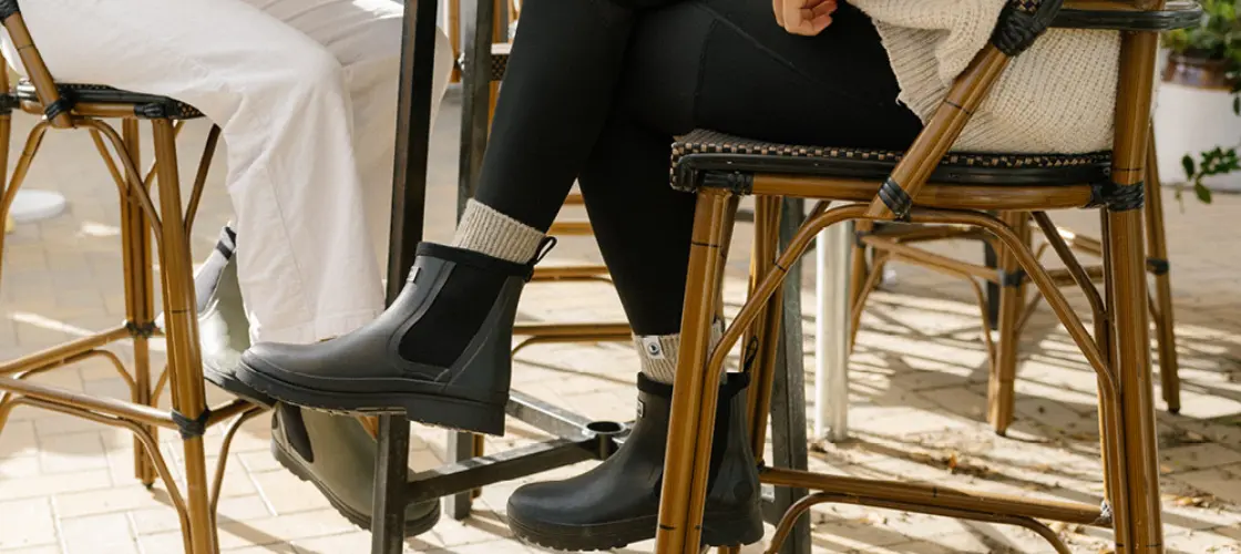Two people sit outdoors on wicker chairs at a table. Both wear black ankle boots; one wears white pants, the other wears black leggings and a cream sweater. Sunlight highlights fallen leaves on the brick floor.