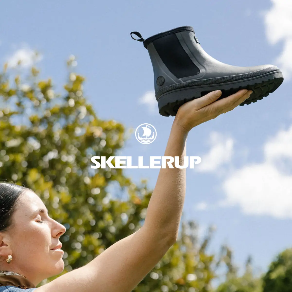 A person holds up a single black boot towards the sky with one hand. The background shows green trees and a clear blue sky. The word SKELLERUP and a logo are centered in the image.