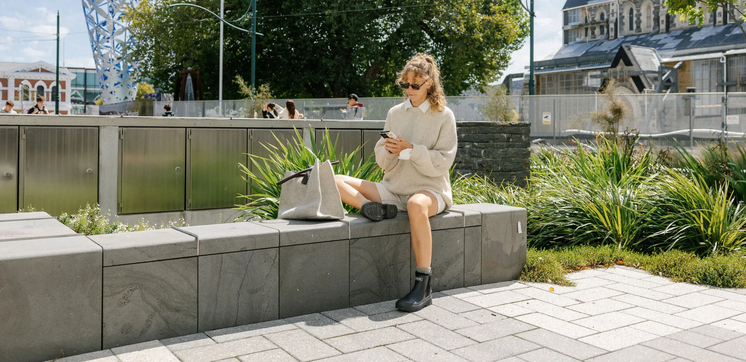 A woman in a beige sweater and boots sits on a stone bench outdoors, looking at her phone. A large tote bag is beside her. Green plants and modern buildings are in the background.