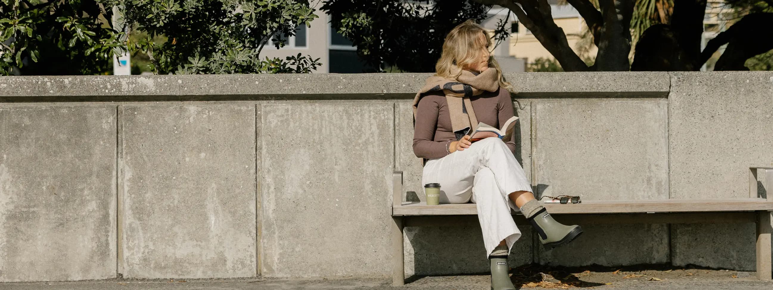 A person sits on a wooden bench outdoors, wearing boots, light pants, and a brown sweater with a scarf. They are reading a book and have a mug and sunglasses next to them. Trees and a concrete wall are in the background.