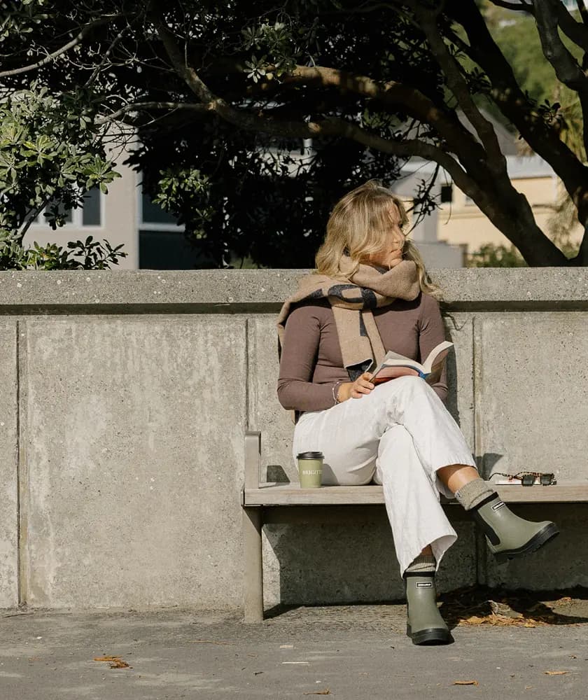 A woman sits on an outdoor bench, wearing white pants, a brown top, green boots, and sunglasses. She holds a notebook, with a coffee cup beside her, and looks to the side under the shade of nearby trees.